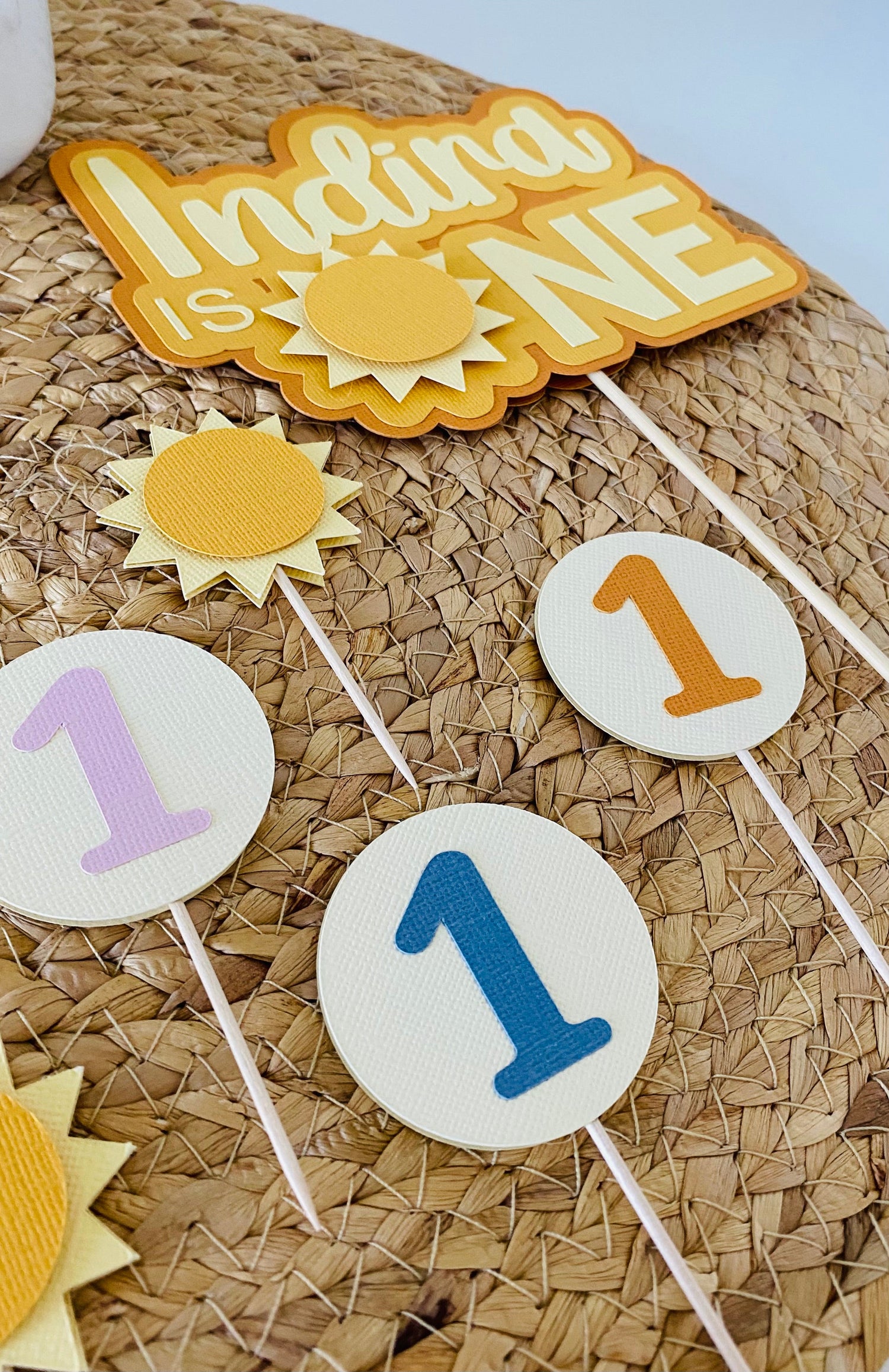Flat lay of First Trip Around the Sun birthday party decor including a cake topper and cupcake toppers in sunshine theme with warm, pastel tones.
