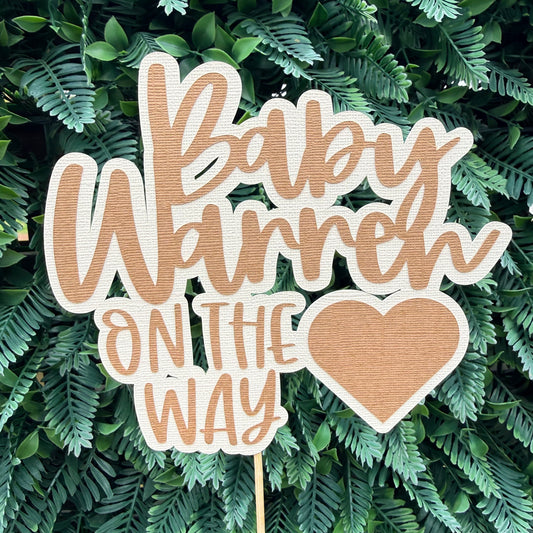Wooden baby shower sign with 'Baby Warren On The Way' text on a green leafy background