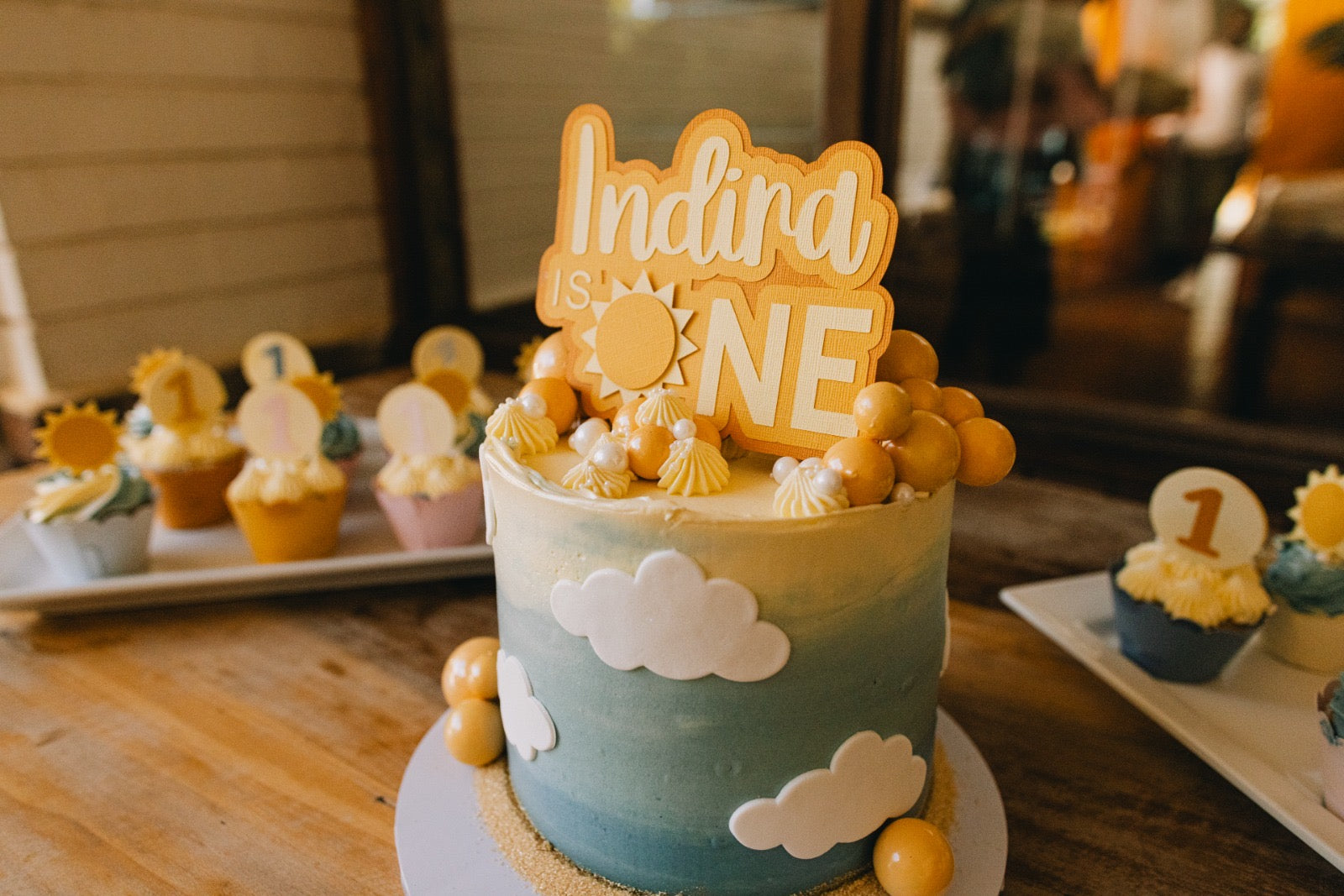 Birthday cake with 'Indira is ONE' topper on a wooden table