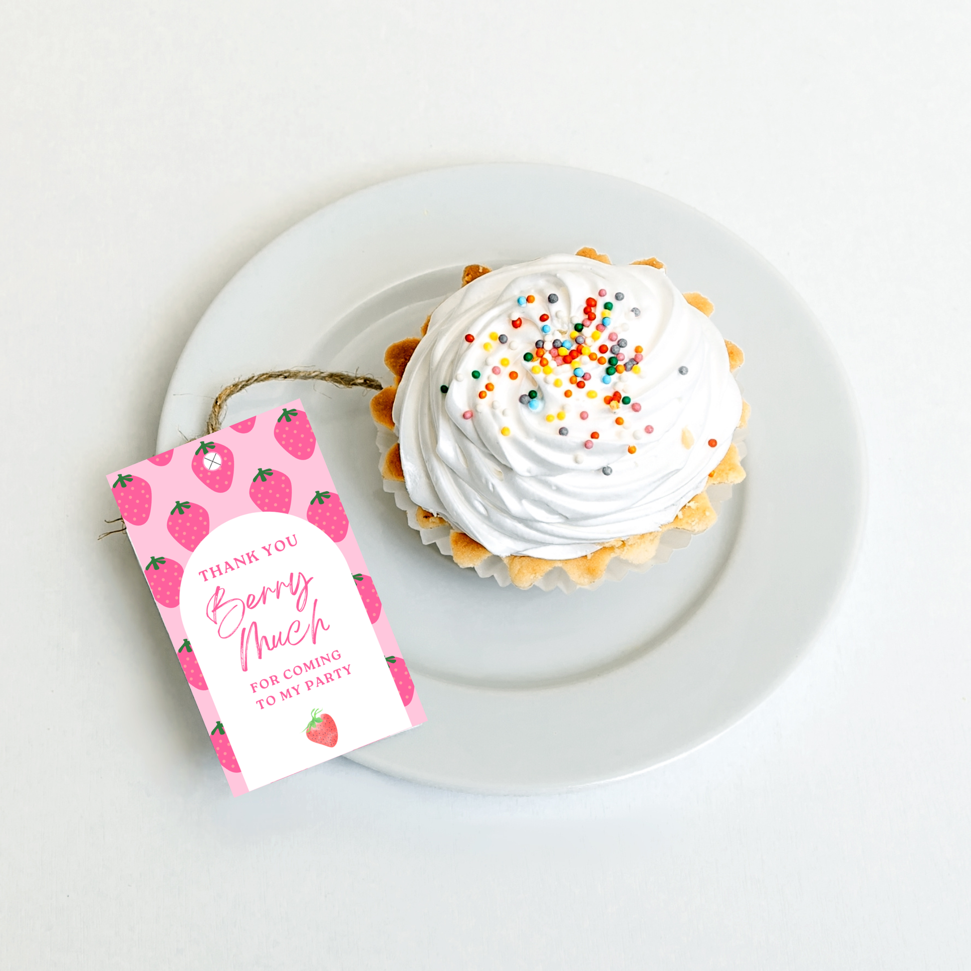 Cupcake with white frosting and colorful sprinkles on a white plate, accompanied by a pink 'Thank You' tag with strawberry design.