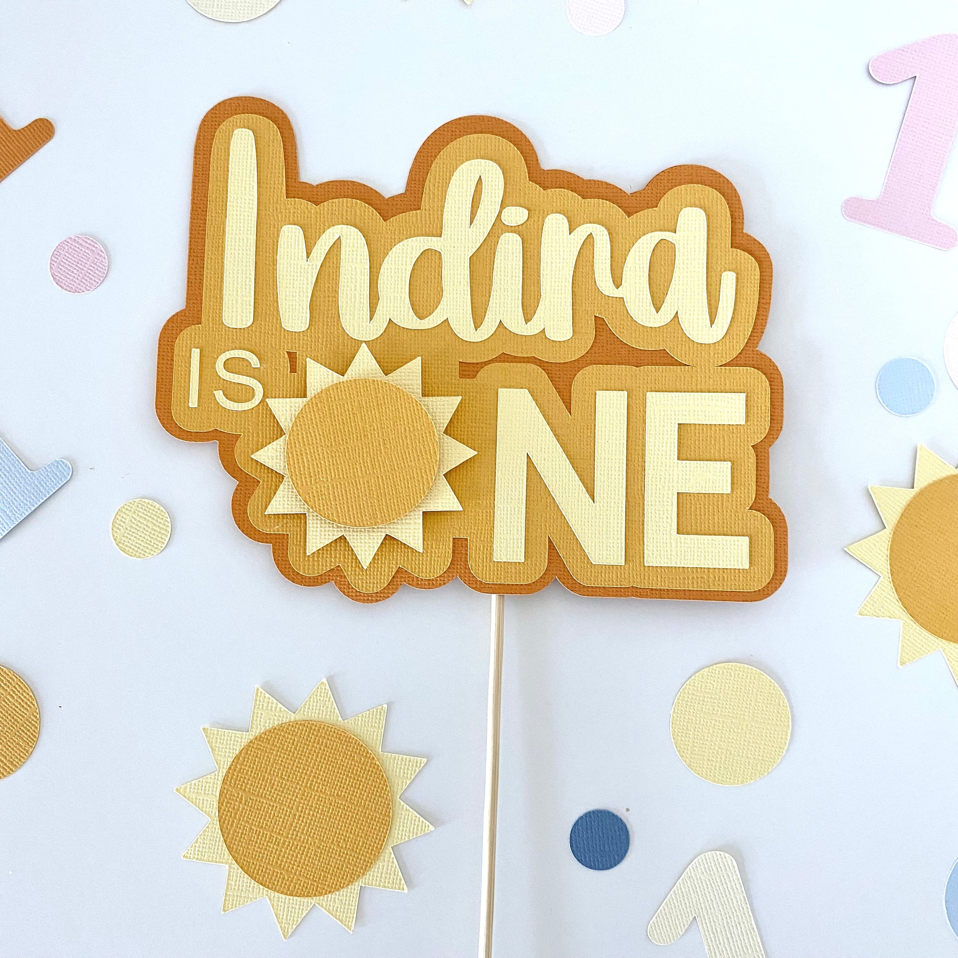 Party decoration with 'Indira is ONE' text on a stick against a light blue background with sun and number decorations.