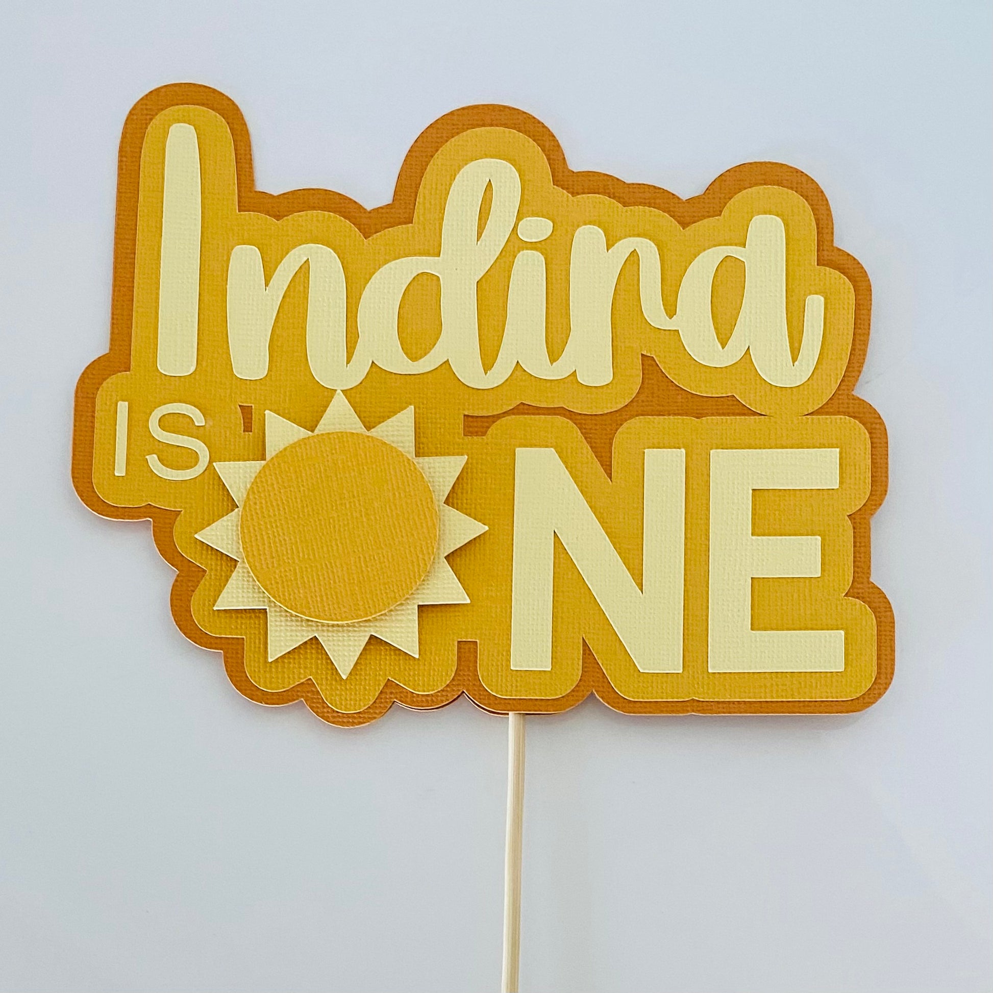Decorative cake topper with 'Indira is ONE' text on a white background
